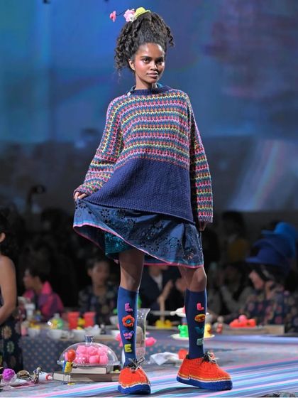 This playful runway look features a high, textured ponytail with small floral accessories. The style was designed to have movement and a youthful feel, matching the colorful knit sweater and patterned socks.