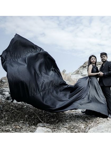 A powerful wide shot of a couple in a rugged, rocky setting, with the black fabric of her trail gown caught in the wind, creating a cinematic effect.