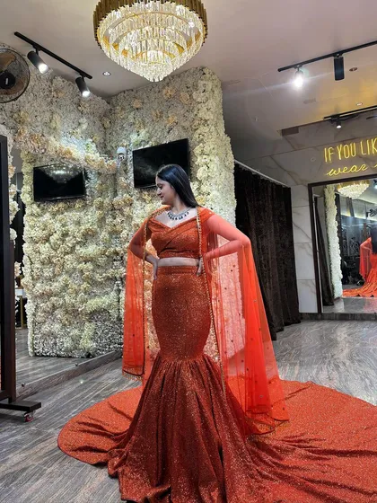 A full-length view of the orange sequined mermaid lehenga. The off-shoulder blouse and cape-style sleeves perfectly complement the figure-hugging silhouette and dramatic trail.