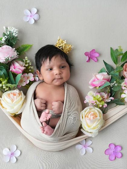 A newborn with a tiny crown peeks out from a cozy swaddle, resting in a fabric hammock adorned with flowers. This shot captures a quiet, awake moment with a touch of royal charm.