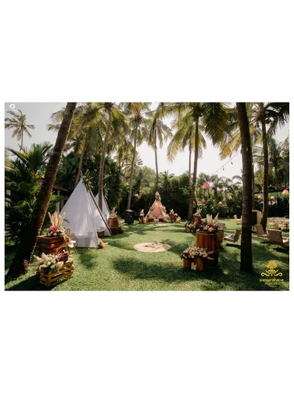 A wide view of a beautiful boho Mehendi setup in Goa, with teepee tents and rustic seating arranged on a lush green lawn under palm trees.