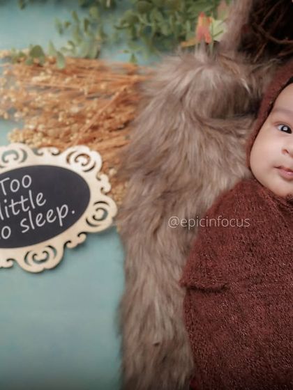 An awake baby peeking out from a warm brown wrap, with a cute sign that reads "Too little to sleep." We capture all the moods of your newborn.
