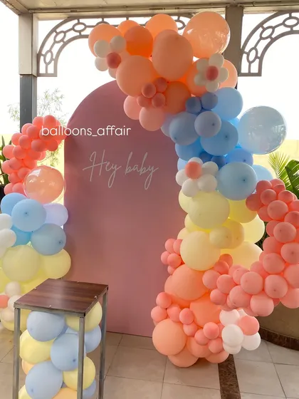 A vibrant pastel rainbow balloon arch against a pink backdrop for a 'Hey Baby' themed celebration.