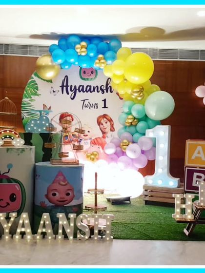 A Cocomelon-themed first birthday with a faux grass floor. The decor includes the iconic yellow bus, character cutouts, and an 'AYAANSH' marquee.