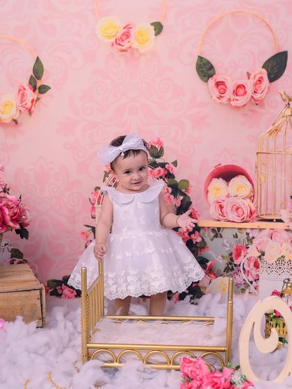 A sweet baby girl in a white dress, standing by a miniature golden bed in our pink floral princess setup. This theme is perfect for creating a fairytale moment.