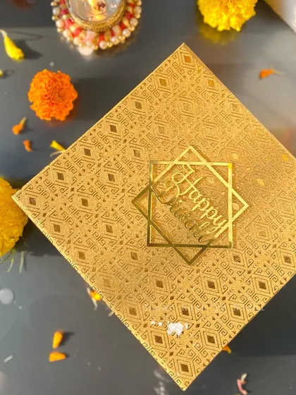 The elegant gold packaging for 'The Sweet Box' for Diwali, surrounded by festive marigold flowers.