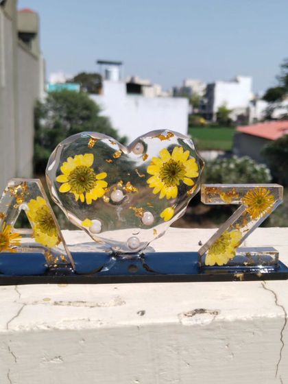 The resin initial frame shown outdoors, where the natural light highlights the clarity of the resin and the vibrant color of the preserved flowers inside.