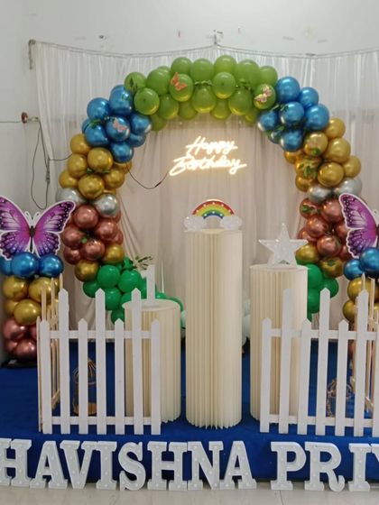 A butterfly garden theme featuring a multi-colored balloon arch against a simple white curtain. The setup includes a neon sign, butterfly cutouts, and pedestals for cake and props.