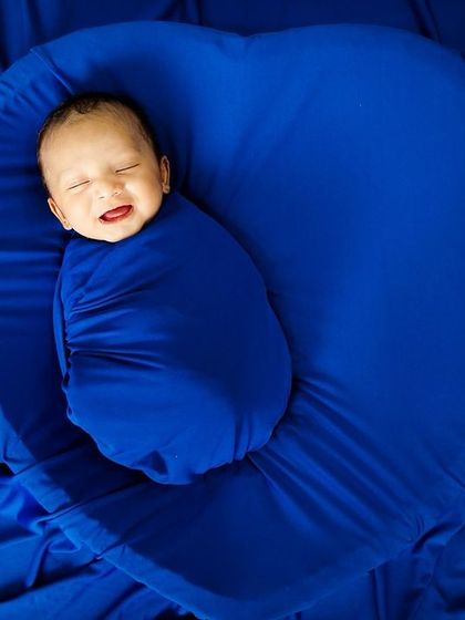 A happy, smiling newborn swaddled snugly in a vibrant blue wrap, nestled in a heart-shaped prop. Capturing these fleeting expressions is my favorite part.