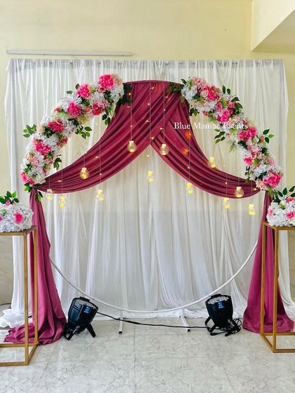 A beautiful Roka ceremony backdrop with a circular frame, wine-colored drapes, and pink and white florals. The hanging glass globes with candles add a magical touch.