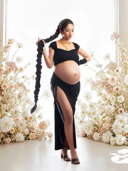 A confident and modern maternity portrait. The client's bold pose, chic black outfit, and long braided hair create a powerful, high-fashion image, beautifully framed by soft flowers.