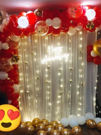 A classic anniversary backdrop with a white curtain and fairy lights. The setup is framed by a red, white, and gold balloon arch and a 'Happy Anniversary' neon sign.