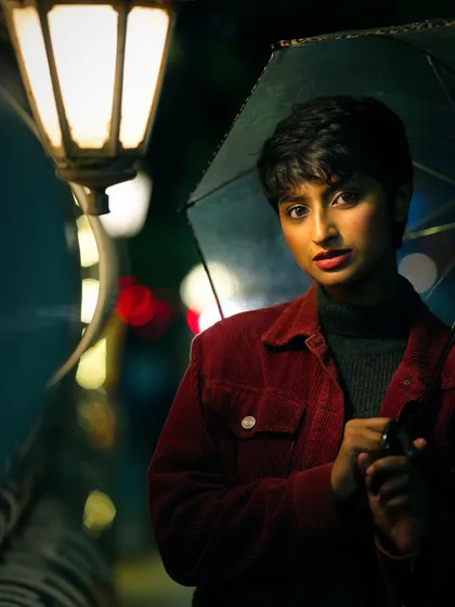 A moody, cinematic portrait of a woman with short hair holding an umbrella under a glowing streetlamp at night. The rich colors and soft focus on the background city lights create a story-driven atmosphere.