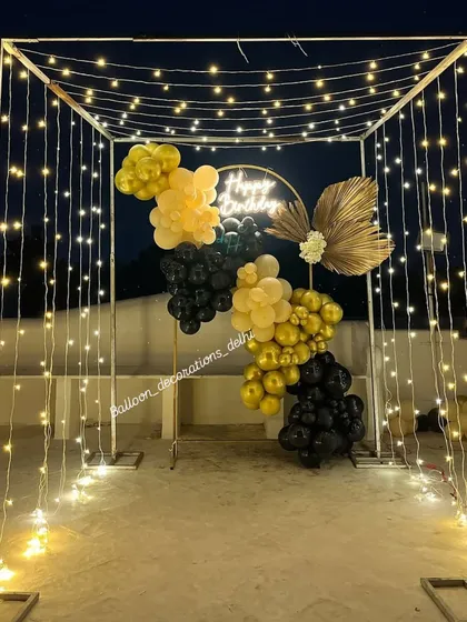 We can transform any outdoor space into a romantic venue. This birthday terrace setup features a beautiful canopy of fairy lights creating a magical tunnel effect, with a stylish black and gold balloon arrangement and a neon sign.