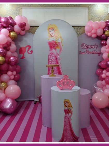 A pretty-in-pink princess-themed second birthday. The setup features an Aurora backdrop, a pink and gold balloon arch, and themed plinths.
