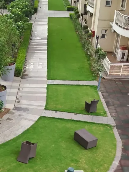 An aerial view of a long, narrow green space in a residential complex. The design provides a green belt between buildings, with simple seating for residents to enjoy.