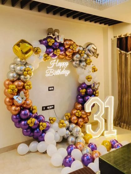 A glamorous 31st birthday decoration for a home party. The setup includes a balloon ring with purple, rose gold, and silver chrome balloons, a neon sign, and large light-up marquee numbers.