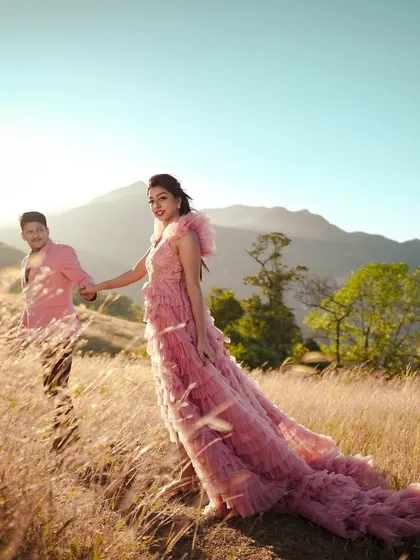 A couple holds hands while walking through a sun-drenched, grassy hillside during their pre-wedding shoot. The bride's flowing pink gown adds a touch of romance to the natural, golden-hour setting.