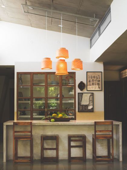 A slightly different angle of the 'Aamra' kitchen, focusing on the cluster of terracotta pendant lights. These lights add warmth and a soft glow to the concrete and wood material palette, enhancing the room's inviting feel.