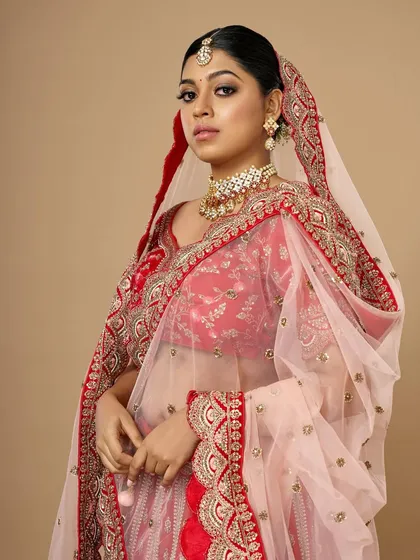 A full-length view of a stunning bride in a red and pink lehenga. The makeup is designed to be impactful from a distance, with defined eyes and a radiant complexion.