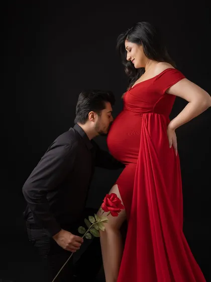 A truly romantic moment from a maternity session. The father-to-be kneels to gently kiss his partner's belly, holding a single red rose. This dramatic studio portrait with a black background highlights their love and excitement.