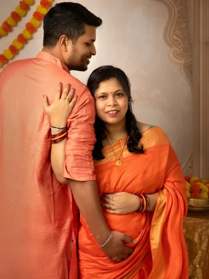 A loving embrace from behind, with the mother-to-be looking warmly at the camera. The vibrant orange and teal sarees, along with the marigold garlands, create a festive and joyful atmosphere.