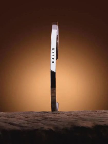 A silver cuff bracelet engraved with 'KARAN', standing vertically on a wooden surface. This artistic shot emphasizes the bracelet's solid construction and sharp engraving.