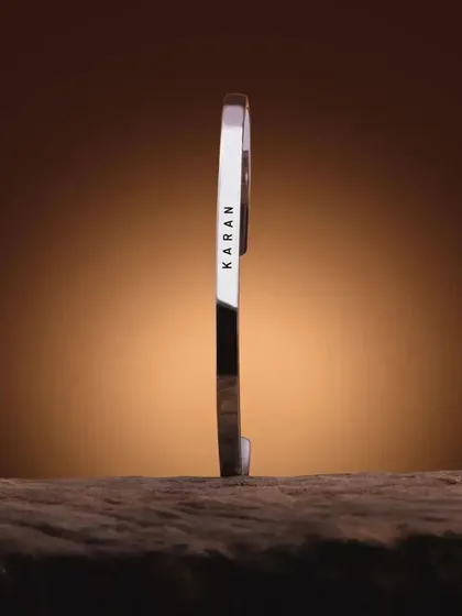 A silver cuff bracelet engraved with 'KARAN', standing vertically on a wooden surface. This artistic shot emphasizes the bracelet's solid construction and sharp engraving.
