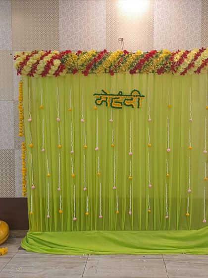 A minimalist Mehandi backdrop with a light green drape and hanging floral buds. The "Mehandi" sign is made of flowers, and the decor is simple yet elegant for an intimate function.