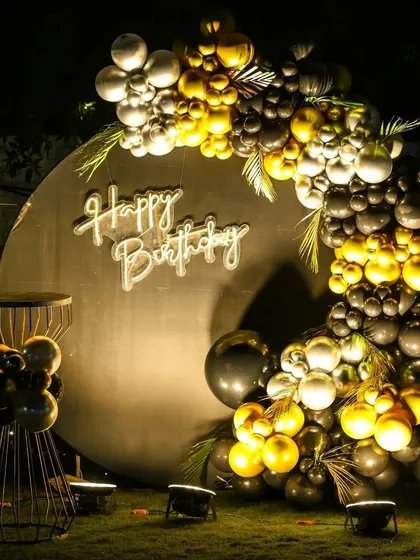 An elegant outdoor birthday setup perfect for an evening celebration. The decor features a large circular backdrop, a "Happy Birthday" neon sign, and a garland of metallic gold and silver balloons.