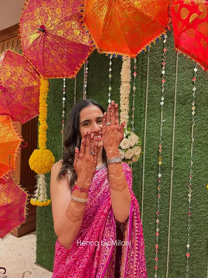 A happy bride showing off her 24-hour henna stain. The deep color looks beautiful against her vibrant pink saree and the festive decor.