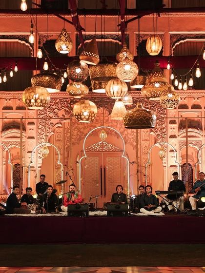 The sangeet stage in its full glory, with the live band performing. The peekaboo arches of the backdrop were designed to complement, not compete with, the stunning palace facade behind it, creating layers of architectural beauty.