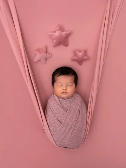Simple, pure, and angelic. This newborn is swaddled snugly in a pink wrap, suspended in a soft sling against a matching backdrop with plush stars, creating a minimalist and dreamy portrait.