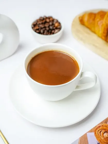 A cup of coffee served with a croissant and a small bowl of coffee beans. The clean white props and background create a sophisticated and minimalist cafe aesthetic.