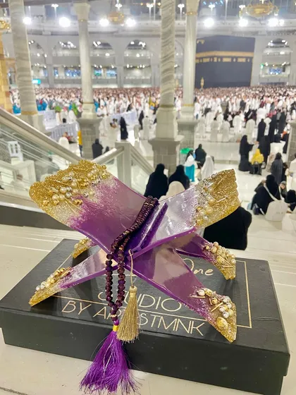 This striking Quran holder is made with a vibrant purple and gold resin design. It comes with a matching crystal tasbeeh and is shown here at the holy city of Makkah, adding to its spiritual significance.