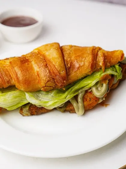A savory croissant sandwich with a green pesto-style sauce and fresh lettuce. The clean, bright photography style makes it look fresh and appealing for a lunch menu.