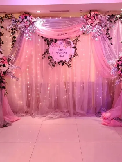 I design elegant backdrops for special occasions like Women's Day. This setup features soft pink draping, twinkling fairy lights, and beautiful floral arrangements, creating a sophisticated and celebratory atmosphere.