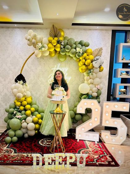 A beautiful olive green and gold theme for Deepu's 25th birthday. The circular balloon arch, "Happy Birthday" neon sign, and large marquee numbers create an elegant and trendy party vibe.