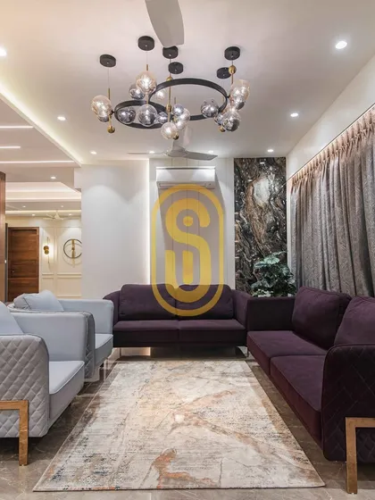 A view of a luxurious living room we designed, featuring a plush purple and grey sofa combination. The space is anchored by a modern statement chandelier and gold accents, creating an atmosphere of sophisticated comfort perfect for entertaining.