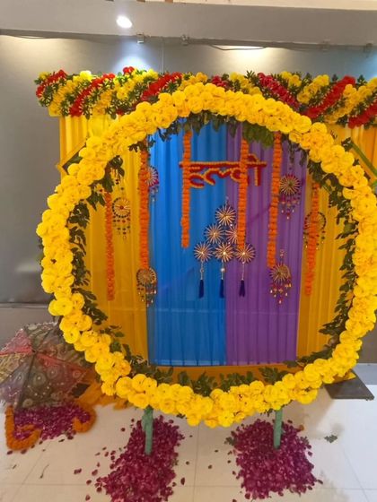 A colorful Haldi setup with a circular marigold backdrop. The decor includes a small stool for the ceremony, Rajasthani umbrellas, and a floor scattered with rose petals.