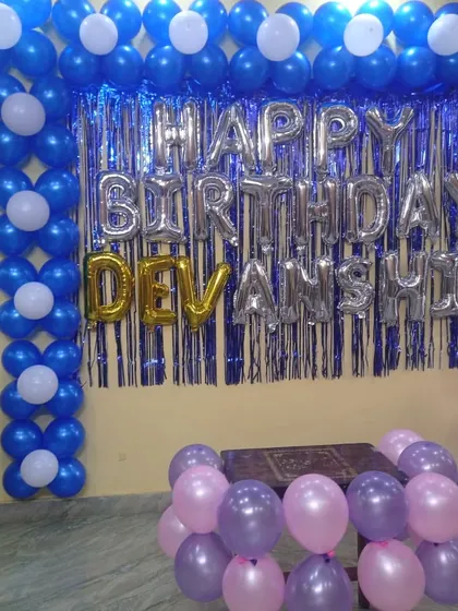 A blue and silver themed birthday decoration. The backdrop features a flower-patterned balloon wall, a shimmer curtain, and the name in foil balloons.