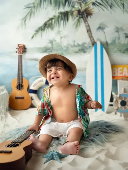 A toddler enjoys a beach-themed studio setup, complete with sand, a surfboard, and a ukulele. A great way to have a beach party without leaving the studio.