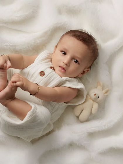 An adorable baby in a white knit romper lies on a soft white blanket, looking at the camera while holding their feet.
