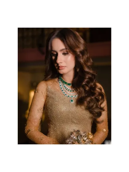 A moment of quiet contemplation. The soft lighting focuses on the bride's expression and the stunning emerald necklace that provides a pop of color against her golden gown, a key styling choice.