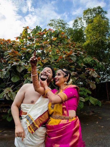 A playful and joyful outdoor portrait. The groom points up at the sky, sharing a laugh with his bride in their vibrant wedding clothes.