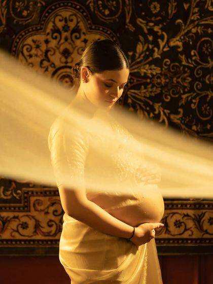 A moment of deep connection. The mother-to-be, draped in a beautiful saree, cradles her belly. The sheer fabric and warm lighting create a soft, ethereal glow, highlighting the sacred bond between mother and child.