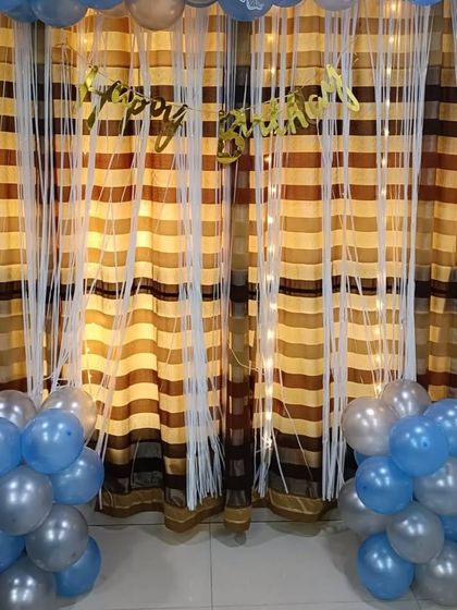 A simple yet beautiful birthday decoration with blue and silver balloon bunches framing a curtain backdrop with fairy lights. This shows how even a small space can be made festive and special with the right balloon arrangement.
