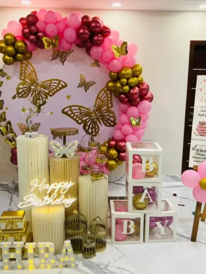 An elegant butterfly-themed first birthday celebration in shades of pink and gold. This decor features a custom backdrop with golden butterflies, a balloon arch, a light-up number '1', and a personalized milestone board for a truly special touch.