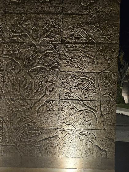 A 'Tree of Life' mural carved into a stone wall, captured at night with uplighting to highlight the detailed texture and craftsmanship.