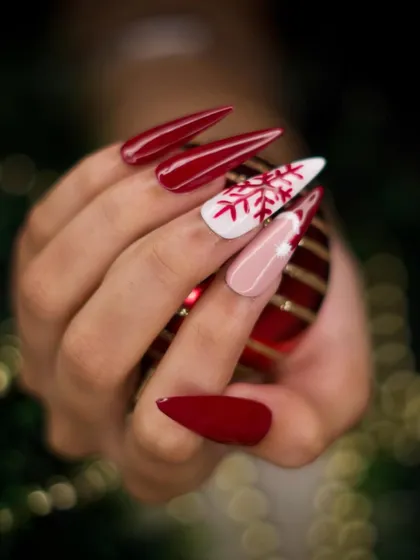 This is a classic holiday manicure featuring sharp, stiletto-shaped extensions. The design pairs a bold, glossy red with accent nails that have a hand-painted snowflake and a subtle Santa hat design.